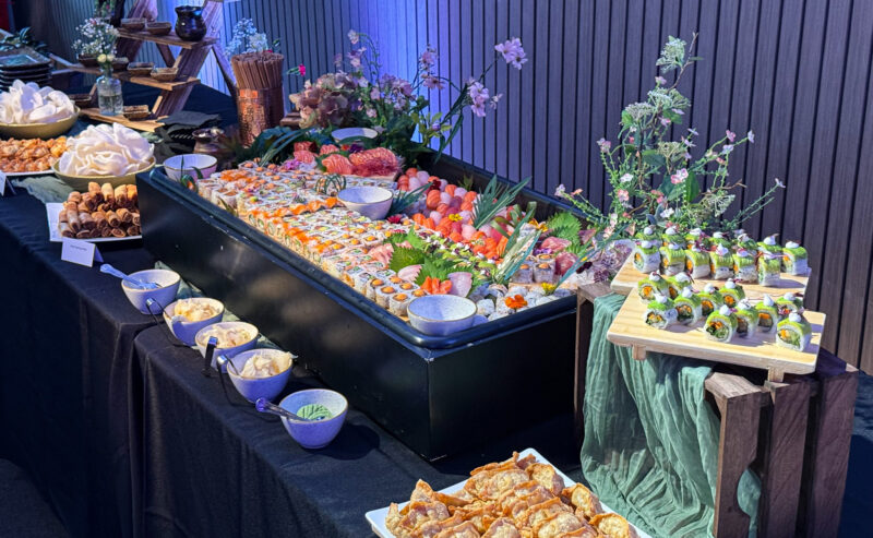 Hurley House - A buffet table displays a large variety of sushi rolls, sashimi, dumplings, and sauces, surrounded by decorative flowers and elegant tableware, set against a wooden wall with blue lighting.