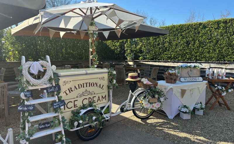 Hurley House - A vintage-style ice cream bike cart, featuring a Traditional Ice Cream sign, is set up outside under a large umbrella in a luxury wedding venue in Berkshire. Adorned with flowers and greenery, its accompanied by a wedding sign, small table, and decorated ladder in the sunny garden setting.