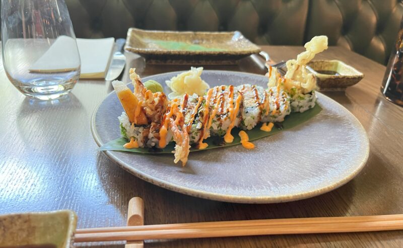 Hurley House - A plate of sushi rolls topped with tempura and orange sauce sits on a table with wooden chopsticks, an empty glass, and small dishes in the background.