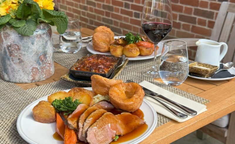 Hurley House - A table set for two with plates of roast beef, Yorkshire pudding, roast potatoes, carrots, and broccoli. There is also red wine, water, a casserole dish, a sauce jug, and a potted flower on the wooden table.