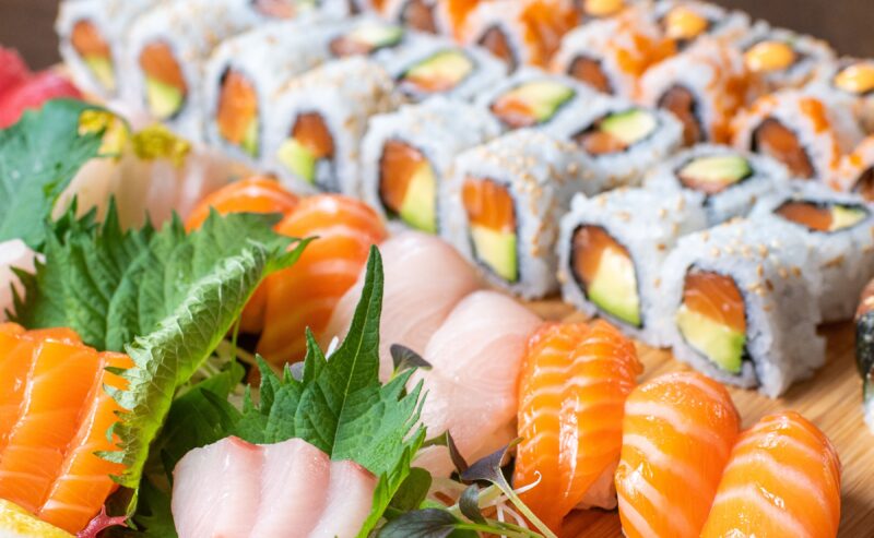 Hurley House - A close-up of assorted sushi, including salmon and tuna sashimi, nigiri, and sushi rolls with rice, avocado, fish, and seaweed, arranged on a wooden board with green shiso leaves.