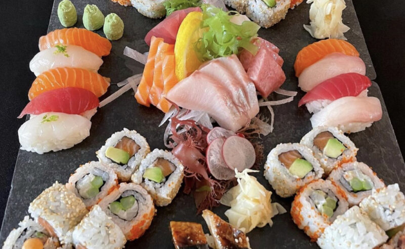 Hurley House - A platter with assorted sushi and sashimi, including various maki rolls, nigiri, sashimi slices, garnishes like wasabi and pickled ginger, arranged artfully on a black slate serving board.