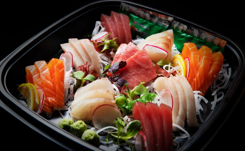 Hurley House - A black tray filled with assorted sashimi, including tuna, salmon, and white fish slices, garnished with radish, lemon, greens, wasabi, and accompanied by soy sauce packets.