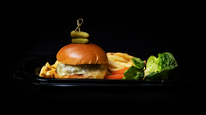 Hurley House - A cheeseburger with a brioche bun topped with two pickles, served with French fries, carrot sticks, and fresh lettuce on a black tray against a black background.