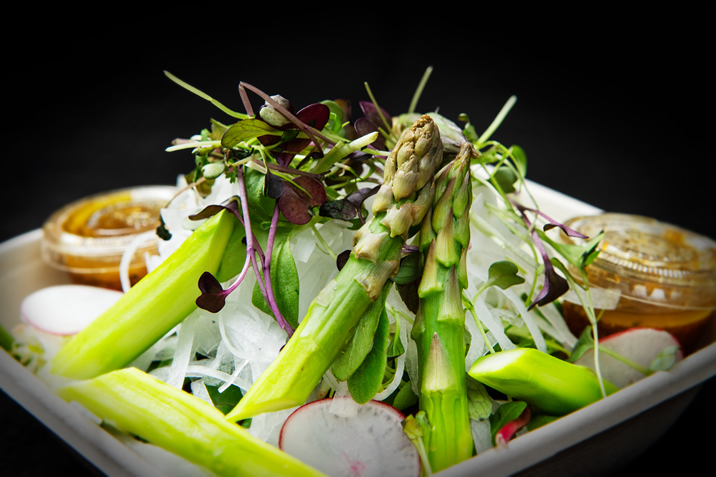 Hurley House - A close-up of a fresh salad featuring asparagus spears, radish slices, microgreens, and shredded vegetables, served in a container with two small cups of dressing in the background.