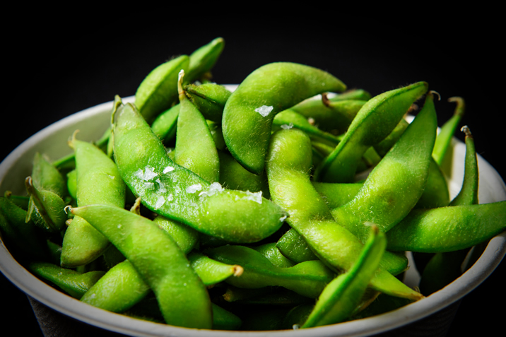 Hurley House - A close-up of a bowl filled with fresh green edamame pods, lightly sprinkled with coarse sea salt, against a dark background.