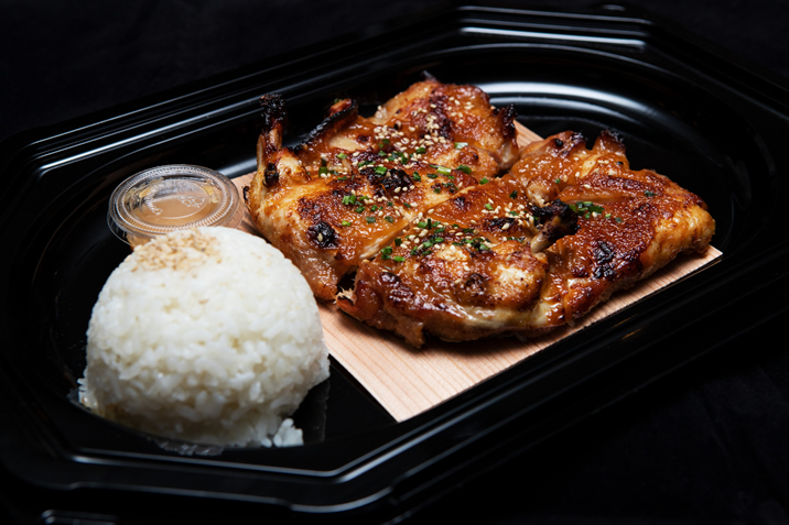 Hurley House - A black tray with grilled chicken garnished with herbs and sesame seeds, a serving of white rice topped with sesame seeds, and a small container of sauce on the side.