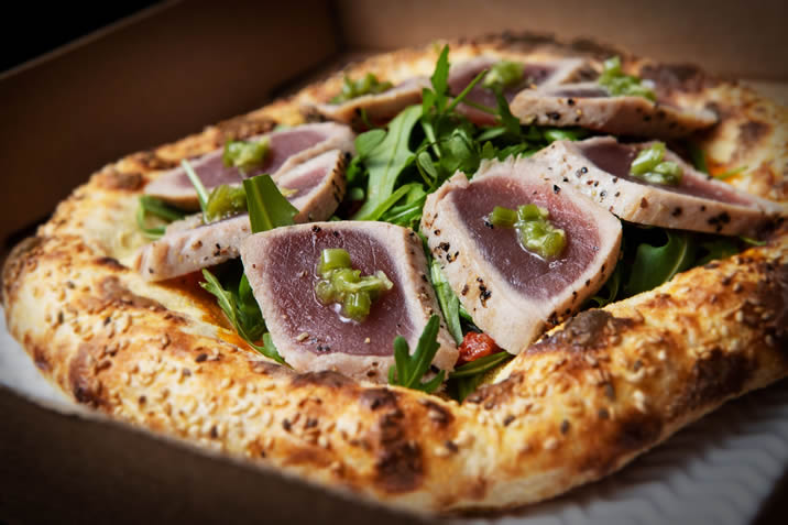 Hurley House - A gourmet pizza topped with seared tuna slices, arugula, diced green peppers, and a thick, golden sesame-seed crust inside a cardboard delivery box.