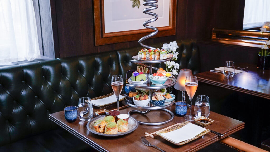 Hurley House - A stylish restaurant table set for two, featuring an elegant spiral stand holding assorted sushi, appetizers, and desserts, with two glasses of sparkling rosé and modern tableware on a dark wooden table.