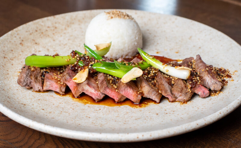 Hurley House - A plate with sliced seared steak topped with green onions, garlic, and sauce, served with a round mound of white rice on a speckled ceramic dish, set on a wooden table.