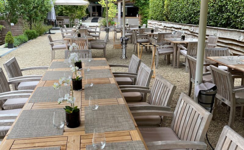 Hurley House - Outdoor dining area with several wooden tables and chairs set up on a gravel patio. Tables have place settings and small potted plants. Large umbrellas provide shade. Green hedges and plants surround the area.