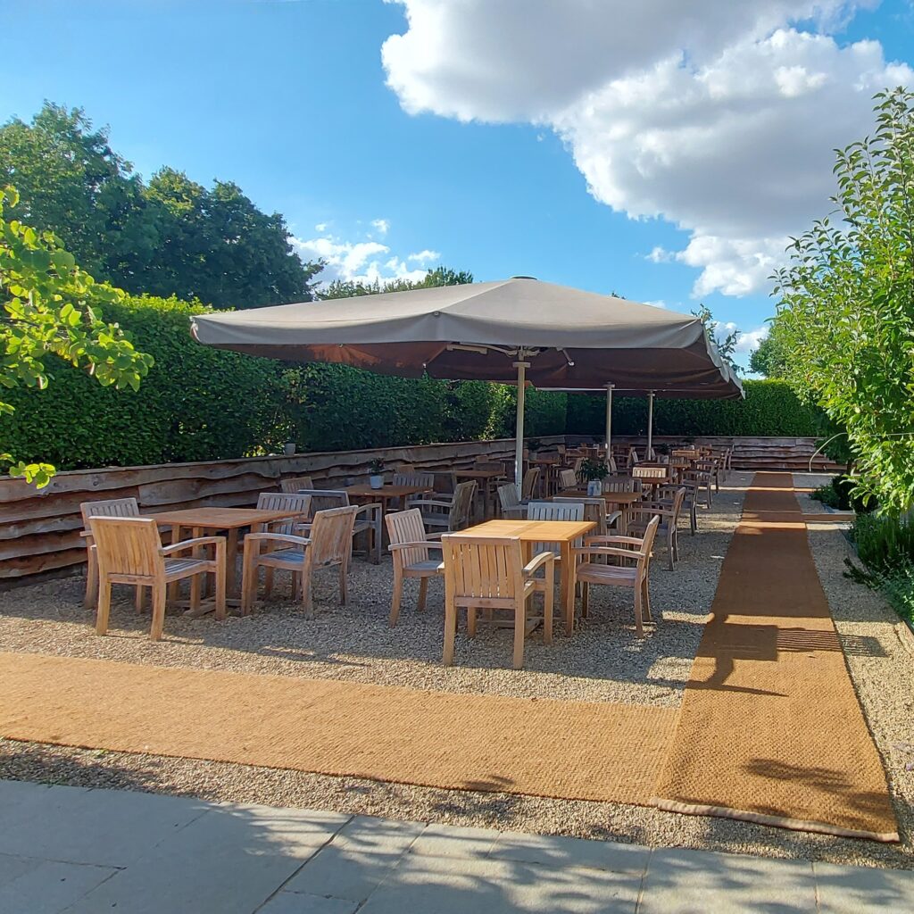 Hurley House - Outdoor seating area with wooden tables and chairs under large umbrellas. The area is surrounded by lush green hedges and trees, with a gravel path leading through it. The sky is partly cloudy with sunshine filtering through.