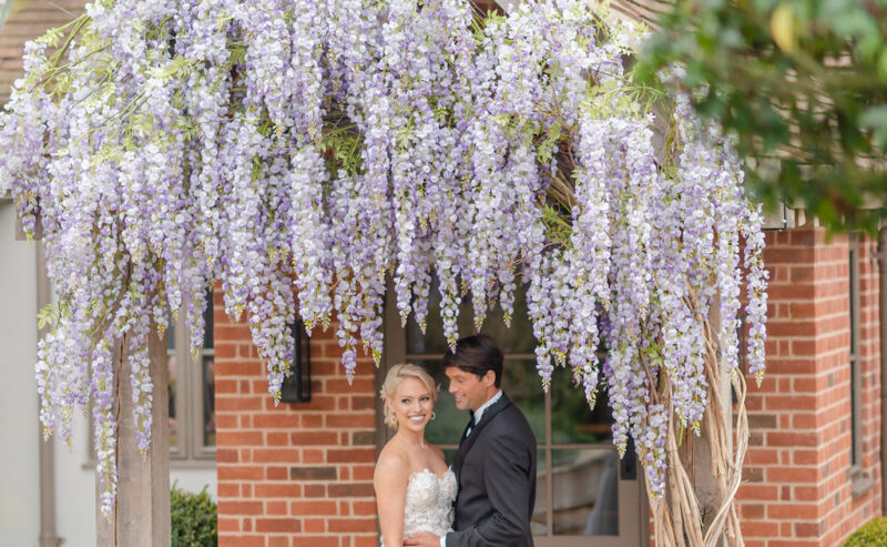 Hurley House - A bride and groom stand beneath a wooden archway adorned with cascading purple wisteria flowers, smiling and embracing outside a brick building surrounded by lush greenery.