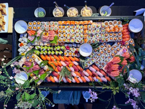 Hurley House - A large platter of assorted sushi, including rolls, nigiri, and sashimi, is artfully arranged on a black tray with decorative flowers around it. Three small bowls with sauces or ginger sit above the platter.