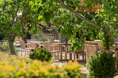 Hurley House - Wooden tables and chairs are arranged outdoors in a garden patio area, surrounded by green trees and shrubs on a sunny day.
