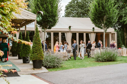 Hurley House - A group of people dressed in formal attire gather and mingle outside a large, elegant marquee tent surrounded by trees and greenery on a sunny day. Tables and chairs are set up on the lawn.