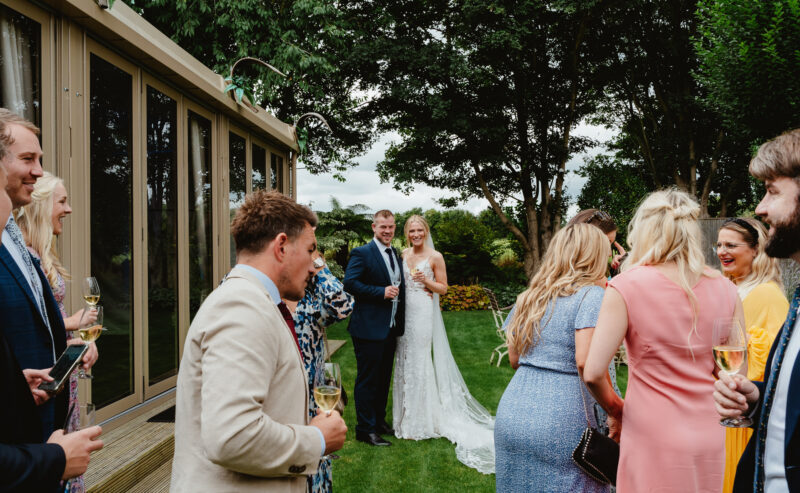 Hurley House - A bride and groom stand together smiling outside as wedding guests in colorful outfits gather around them, holding drinks and chatting on a grassy lawn near a building and trees.