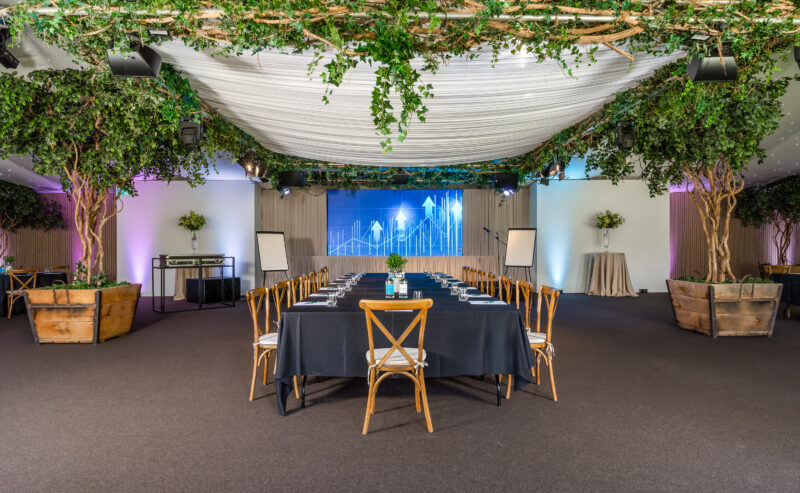 Hurley House - A conference room with a long table covered in a black tablecloth, surrounded by wooden chairs. The ceiling is decorated with draped white fabric and greenery, and potted trees line the room. A screen and flipcharts are at the front.