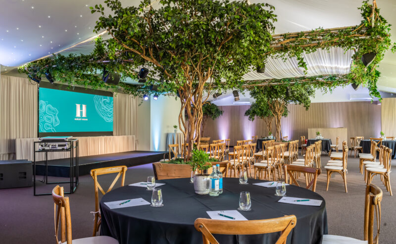 Hurley House - A spacious event room with round tables and wooden chairs, decorated with large indoor trees and greenery. A stage with a screen displaying an “H” logo is set up at the front. Glasses, water bottles, and notepads are on the tables.