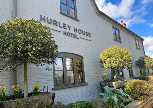 Hurley House - A white brick building with the sign Hurley House Hotel features large windows, potted trees, and yellow flowers along the front, set against a blue sky with scattered clouds.
