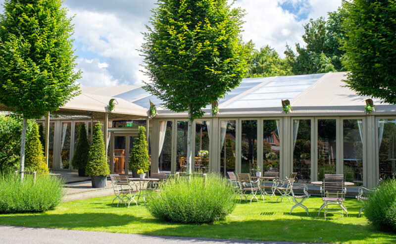 Hurley House - A serene outdoor scene at a luxury wedding venue in Berkshire, featuring a large tented event space surrounded by tall triangular shrubs and trees. Rows of metal chairs and small tables dot the lush green lawn, all under a partly cloudy sky.