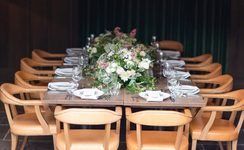 Hurley House - A long dining table is elegantly set with white plates, folded napkins, and glassware at a boutique wedding venue in Berkshire. The centerpiece features lush floral arrangements with green and pink tones. Surrounding the table are ten tan leather chairs.