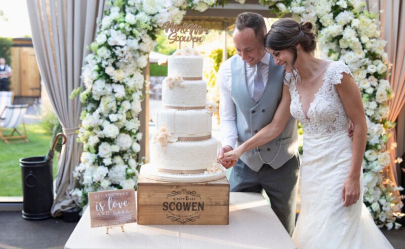 Hurley House - A bride and groom smile while cutting a three-tiered wedding cake adorned with flowers under a floral arch, Mr & Mrs visible above. A wooden sign reads Scowen, all set in a luxury wedding venue in Berkshires lush garden.
