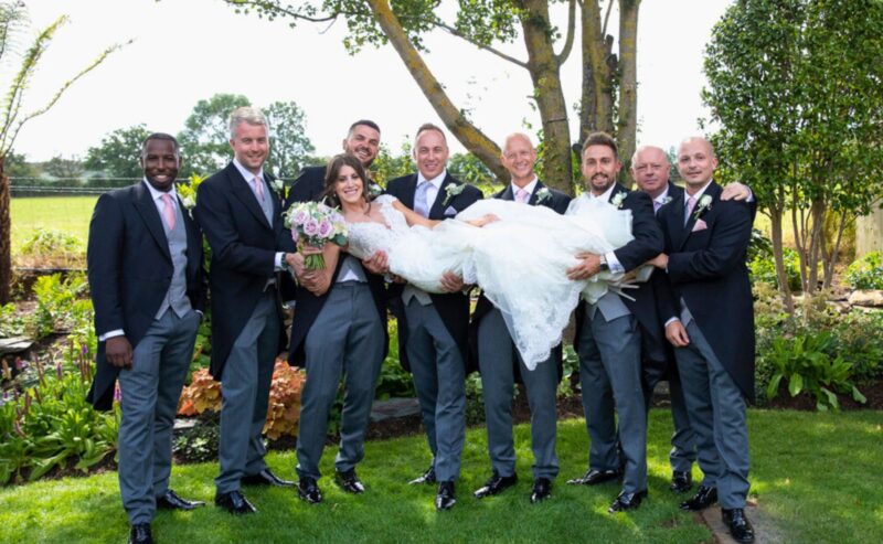 Hurley House - A bride in a white gown is being playfully lifted by a group of seven groomsmen in matching suits at a luxury wedding venue in Berkshire. They are all smiling, surrounded by lush trees and plants, as the sun gently bathes the grassy area where they stand.