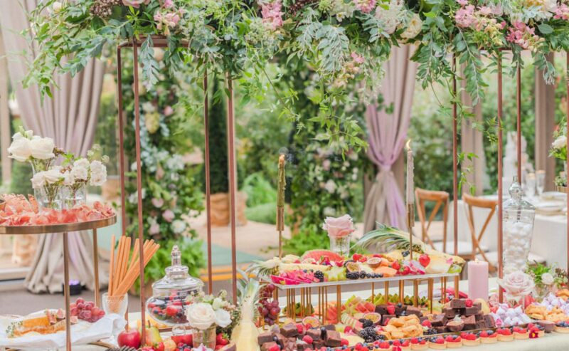 Hurley House - A beautifully arranged charcuterie board graces the wedding buffet at a luxury venue in Berkshire, filled with fruits, cheeses, and meats. Elegant floral decorations overhead feature pink and white roses with greenery, set against the backdrop of soft drapes and a picturesque garden.