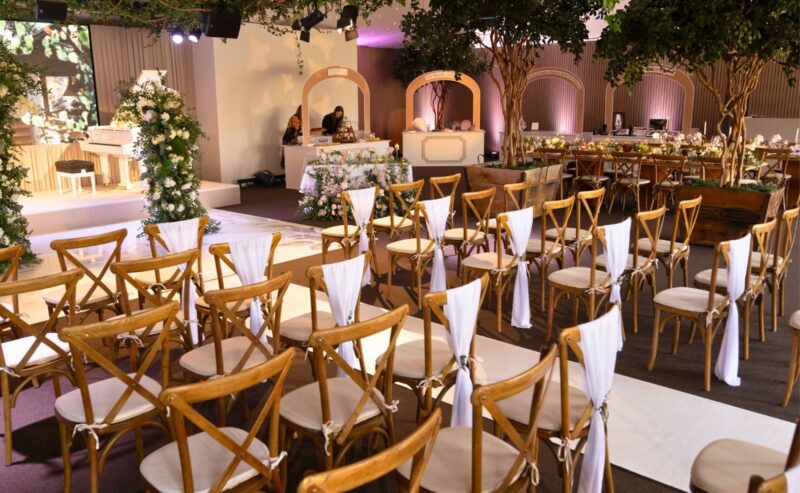 Hurley House - Experience an elegant indoor luxury wedding venue in Berkshire, featuring wooden chairs draped in white fabric. Floral decorations and greenery enhance the space, with a stage showcasing floral backdrops and wedding tables in the background.
