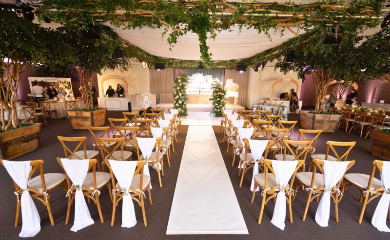 Hurley House - The elegant indoor wedding ceremony setup boasts wooden chairs draped in white fabric along a central aisle. Surrounded by greenery and flowers under a draped ceiling, this luxury wedding venue in Berkshire offers an inviting and romantic atmosphere.