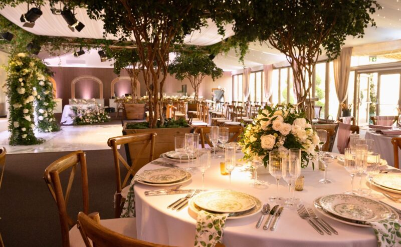 Hurley House - An elegant luxury wedding venue in Berkshire, featuring a reception setup with round tables adorned with floral centerpieces, fine china, and glassware. The room boasts draped ceilings, greenery, and large windows that let natural light illuminate the space.