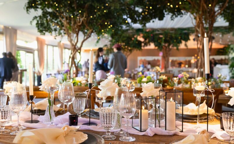Hurley House - Elegant wedding table setting at a luxury wedding venue in Berkshire, featuring crystal glasses, white rose centerpieces, and lit candles in glass holders. Soft lighting and blurred guests in the background create a warm, festive atmosphere under lush greenery and fairy lights.